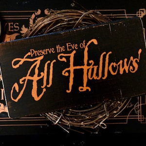 Op de afbeelding: Een zwart houten bord met oranje tekst die "Preserve the Eve of All Hallows" luidt. Het bord ligt op een zwart oppervlak met een bruine krans.
