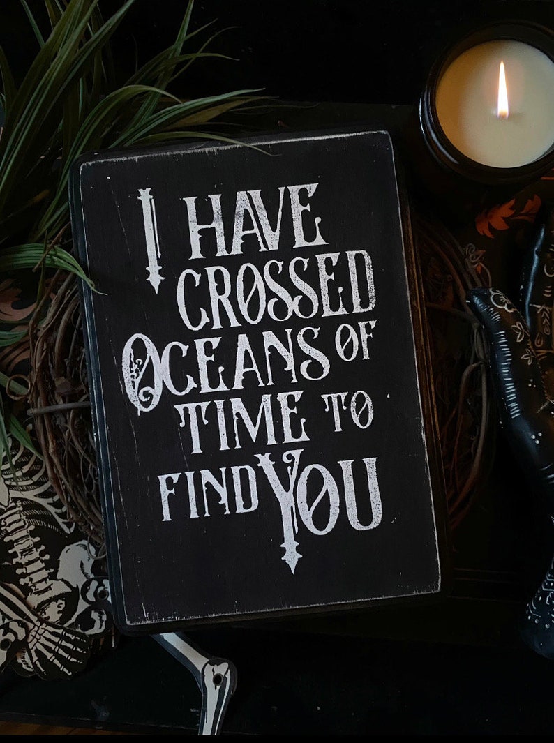 May include: A black wooden sign with white lettering that reads "I HAVE CROSSED OCEANS OF TIME TO FIND YOU." The sign is surrounded by a candle, greenery, and decorative items, creating a gothic or romantic aesthetic.