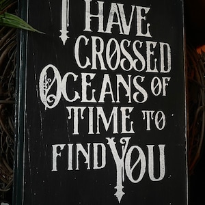 May include: Black wooden sign with white lettering that reads "I HAVE CROSSED OCEANS OF TIME TO FIND YOU." The sign has a distressed, vintage look with visible wood grain and a dark border. The text is in a decorative, serif font.