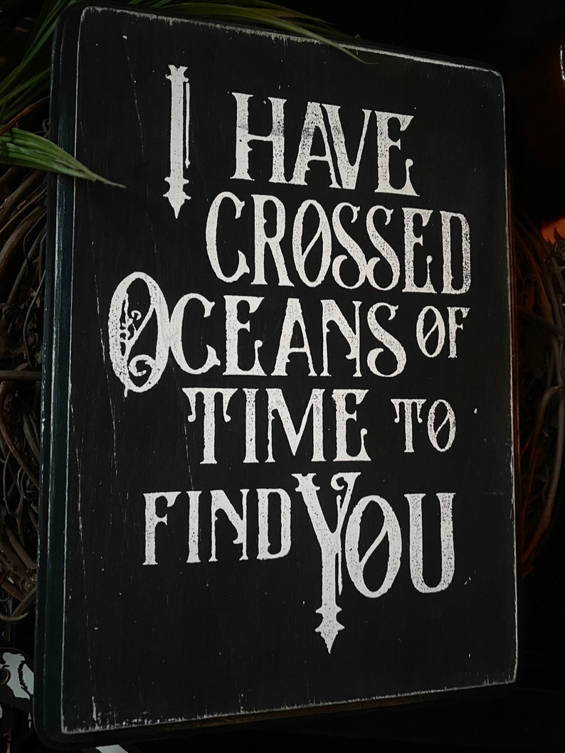 May include: Black wooden sign with white lettering that reads "I HAVE CROSSED OCEANS OF TIME TO FIND YOU." The sign has a distressed, vintage look with visible wood grain and a dark border. The text is in a decorative, serif font.