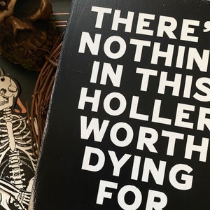 May include: Black wooden sign with white text that reads "There's nothing in this holler worth dying for."