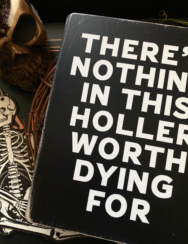 May include: Black wooden sign with white text that reads "There's nothing in this holler worth dying for."