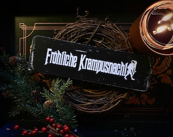 Può includere: Un cartello in legno nero con testo bianco che recita "Fröhliche Krampusnacht!" con una silhouette bianca di una figura di Krampus sul lato destro. Il cartello è appoggiato su una ghirlanda di rami marroni.