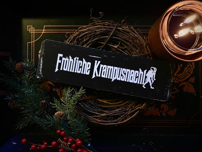 May include: A black wooden sign with white text that reads "Fr&ouml;hliche Krampusnacht!" with a white silhouette of a Krampus figure on the right side. The sign is sitting on a brown twig wreath.