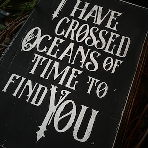 May include: A black wooden sign with white lettering. The text reads "I HAVE CROSSED OCEANS OF TIME TO FIND YOU." The sign has a distressed, vintage look, with visible wood grain and a slightly worn appearance. The background is dark.