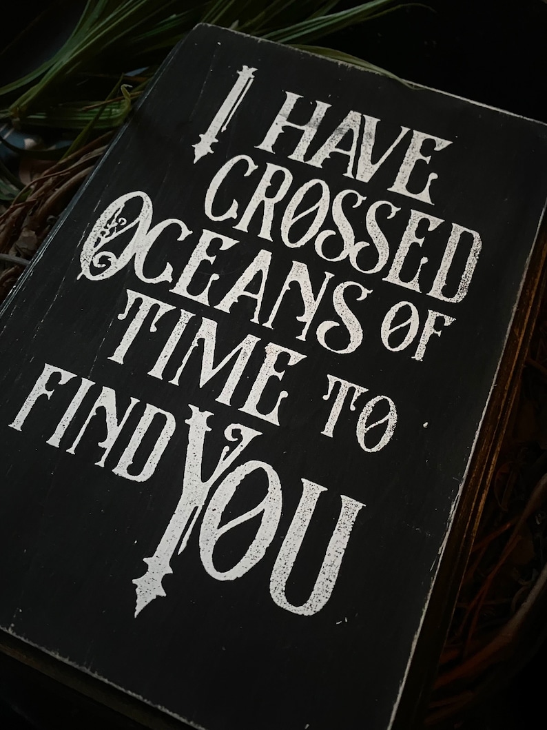 May include: A black wooden sign with white lettering. The text reads "I HAVE CROSSED OCEANS OF TIME TO FIND YOU." The sign has a distressed, vintage look, with visible wood grain and a slightly worn appearance. The background is dark.