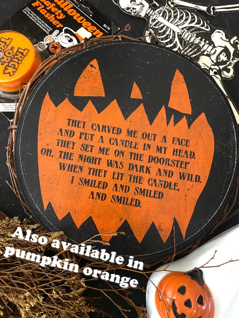 Op de afbeelding: Een ronde, zwarte Halloween-decoratie met een oranje pompoengezichtontwerp en tekst. De tekst luidt: "Ze sneden een gezicht uit... Ik glimlachte en glimlachte en glimlachte." De decoratie is omgeven door een gevlochten krans.