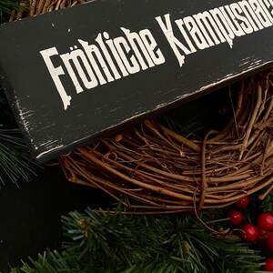 May include: A black wooden sign with white lettering that reads "Fr&ouml;hliche Krampusnacht". The sign is hanging on a brown grapevine wreath with red berries.