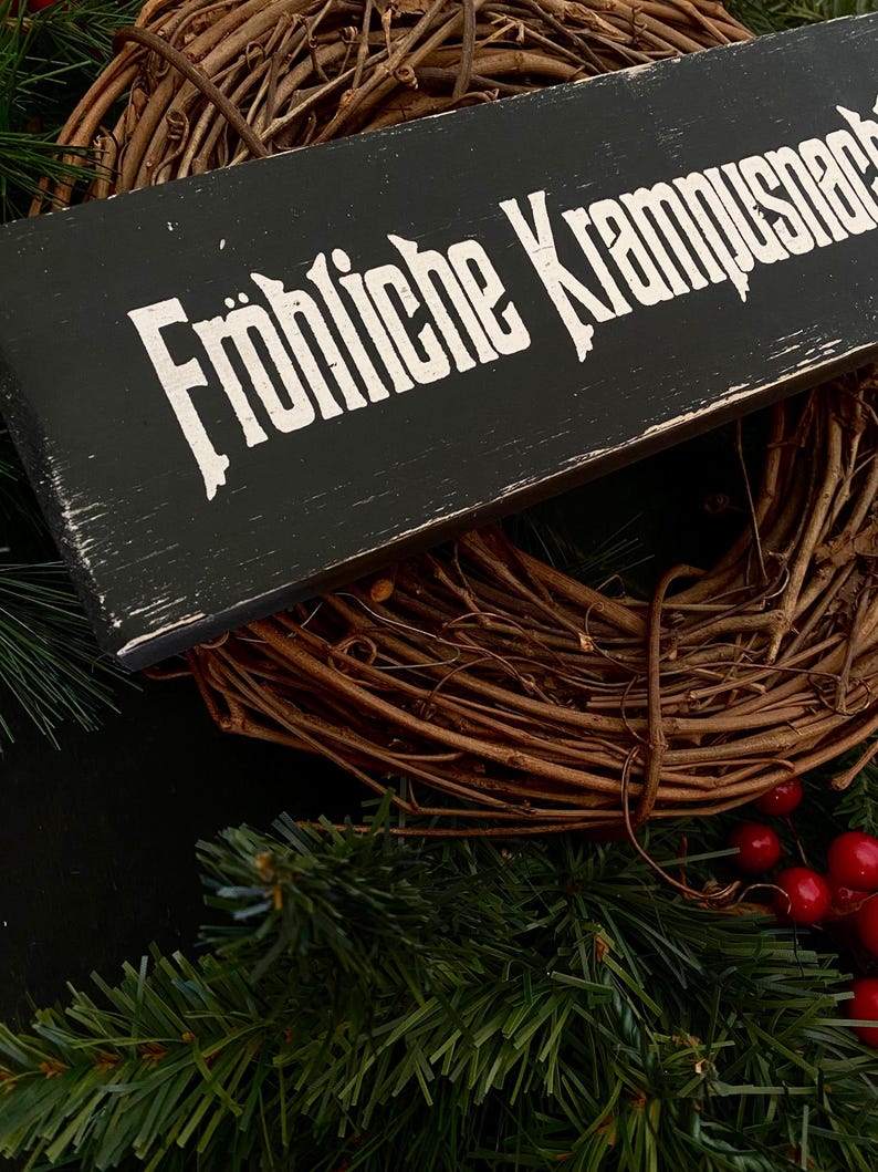 May include: A black wooden sign with white lettering that reads "Fr&ouml;hliche Krampusnacht". The sign is hanging on a brown grapevine wreath with red berries.