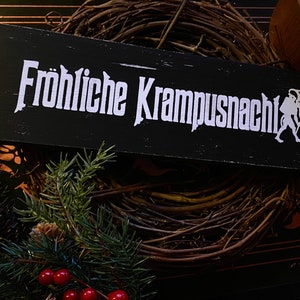 May include: A black wooden sign with white text that reads "Fr&ouml;hliche Krampusnacht" and a white silhouette of a Krampus figure. The sign is sitting on a brown twig wreath.
