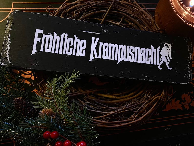 May include: A black wooden sign with white text that reads "Fr&ouml;hliche Krampusnacht" and a white silhouette of a Krampus figure. The sign is sitting on a brown twig wreath.
