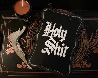 May include: A black wooden sign with the words "Holy Shit" in white gothic lettering. A lit orange candle in a black jar is to the left, and a jawbone is in the foreground. The sign is surrounded by a twig wreath.