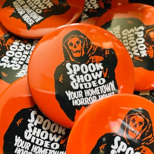 May include: A close-up of several orange round buttons featuring a black and white image of a hooded figure with a skull face. The text on the buttons reads "Spook Show Video Your Hometown Horror House".