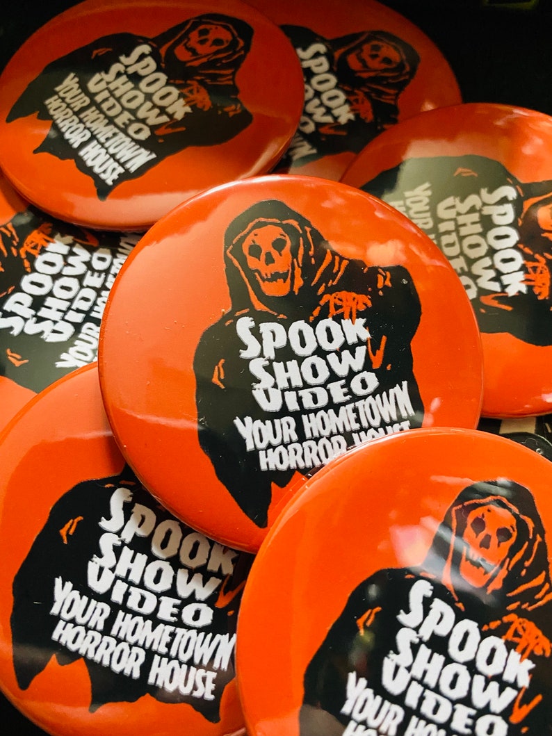May include: A close-up of several orange round buttons featuring a black and white image of a hooded figure with a skull face. The text on the buttons reads "Spook Show Video Your Hometown Horror House".