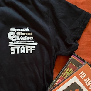 May include: Black t-shirt with white text that reads "Spook Show Video Your Hometown Horror House" and "STAFF".