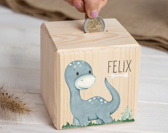 May include: A wooden piggy bank with a blue dinosaur illustration and the name "FELIX" painted on the front. A coin is being inserted into the slot.