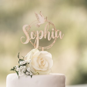 Peut inclure: Un gâteau blanc avec un topper de gâteau en bois représentant une colombe et le nom "Sophia" en écriture cursive. Le gâteau est décoré d'une rose blanche et de petites fleurs blanches.