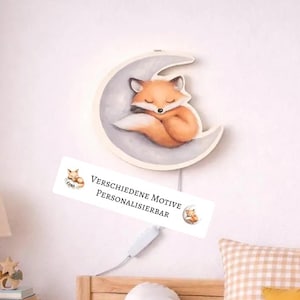 Può includere: Una luce notturna a forma di mezzaluna, con un design di una volpe che dorme. La volpe è arancione e bianca, appoggiata su una luna grigia. La luce ha un cavo bianco e il testo "VERSCHIEDENE MOTIVE PERSONALISIERBAR".