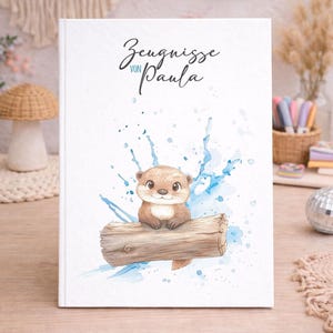 Può includere: Un libro con copertina rigida bianca con il testo "Zeugnisse von Paula" in corsivo. La copertina presenta un'illustrazione ad acquerello di una simpatica lontra seduta su un tronco con schizzi d'acqua blu. Il libro è su un tavolo di legno.