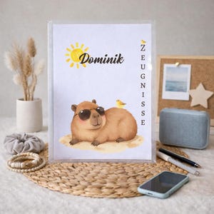 Könnte beinhalten: Eine transparente Kunststoff-Dokumentenmappe mit einer Illustration eines Capybaras mit Sonnenbrille, dem Namen "Dominik" und einer Sonne. Ein kleiner gelber Vogel sitzt auf dem Rücken des Capybaras. Das Wort "ZEUGNISSE" ist vertikal gedruckt.