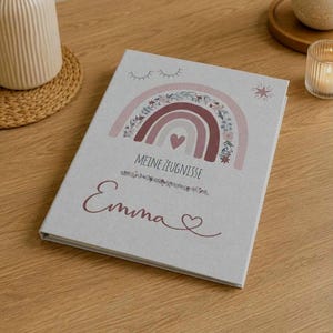 May include: A light gray hardcover book featuring a printed design of a floral rainbow with a heart, closed eyes, and a star. The text reads "MEINE ZEUGNISSE" above the name "Emma" which is written in a cursive script with a small heart at the end.