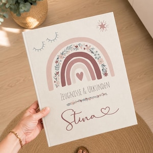 May include: A white hardcover book featuring a printed design of a rainbow with floral accents, a small heart, closed eyes, and a star. The text reads ZEUGNISSE & URKUNDEN and the name Stina in a cursive script with a small heart symbol at the end.