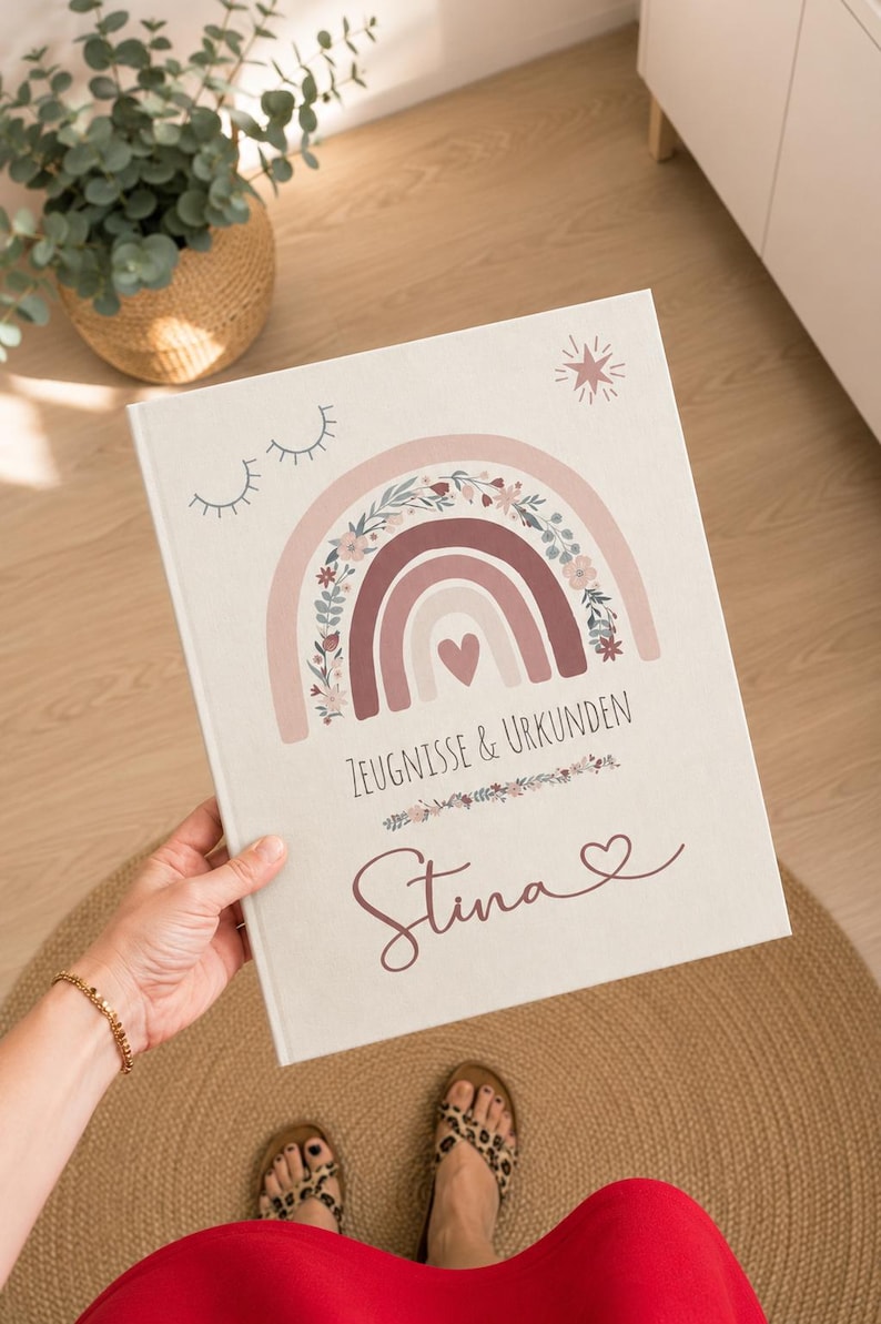 May include: A white hardcover book featuring a printed design of a rainbow with floral accents, a small heart, closed eyes, and a star. The text reads ZEUGNISSE & URKUNDEN and the name Stina in a cursive script with a small heart symbol at the end.