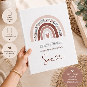 May include: A white binder for certificates and documents featuring a boho-style rainbow illustration with floral accents in muted earth tones. The text reads "ZEUGNISSE & URKUNDEN" and the name "Soe" with a heart symbol below the rainbow.