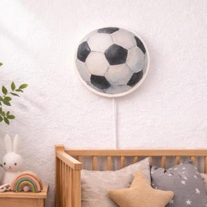 Könnte beinhalten: Eine runde Wandleuchte in Form eines Fußballs, mit weißen und schwarzen Fünfecken. Sie ist an einer weißen Wand befestigt, mit einem weißen Kabel, das nach unten hängt. Das Licht befindet sich über einem hölzernen Bettrahmen.