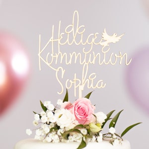 Könnte beinhalten: Ein weißer Kuchen mit einem Holzaufsatz, auf dem "Heilige Kommunion Sophia" mit einem Taubenmotiv steht. Der Kuchen ist mit weißen und rosa Blumen verziert.