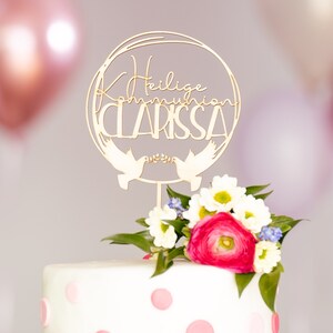 Può includere: Un topper per torta in legno dorato con il testo "Heilige Kommunion Clarissa" e due colombe che tengono un ramo con foglie. Il topper è su una torta bianca con pois rosa e fiori rosa e bianchi.