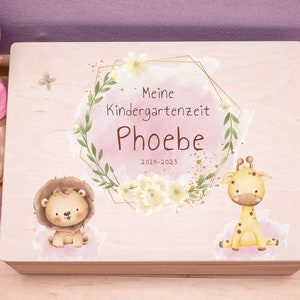 Può includere: Una scatola di legno per ricordi con un design floreale rosa e oro. La scatola ha il testo "Meine Kindergartenzeit Phoebe 2019-2023" sul davanti. Ci sono illustrazioni di un leone e una giraffa sulla scatola.