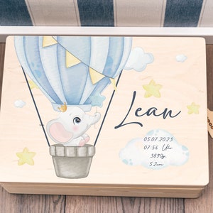 Peut inclure: Une boîte à souvenirs en bois avec un motif de ballon à air chaud bleu clair et blanc. Un éléphant est assis dans le ballon. La boîte est décorée d'étoiles jaunes et le nom "Lean" est écrit sur le devant. La date "05.07.2023", l'heure "07:56 Uhr", le poids "3890g" et la longueur "52cm" sont également inscrits sur la boîte.