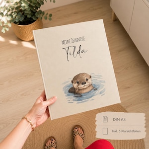 Può includere: Una cartella portadocumenti A4 color beige con il testo "MEINE ZEUGNISSE" e il nome "Tilda" in corsivo nero, un'illustrazione ad acquerello di una lontra marrone che nuota nell'acqua blu, inclusi cinque fogli protettivi trasparenti.