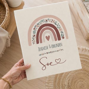 May include: A rectangular off-white binder featuring a printed design of a rainbow with floral accents and a central heart. The text "ZEUGNISSE & URKUNDEN" is printed above the name "Soe" followed by a small heart symbol in dark red ink.