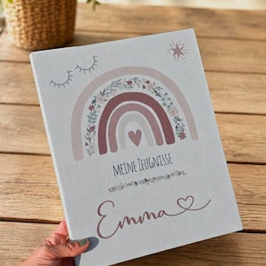 May include: A light gray rectangular folder featuring a printed design of a muted pink and beige rainbow with floral accents, a small heart, and closed eyes. The text reads "MEINE ZEUGNISSE" above the name "Emma" with a small heart symbol.