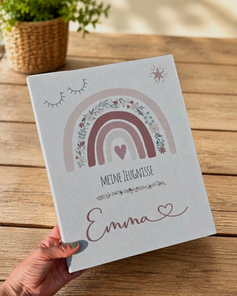 May include: A light gray rectangular folder featuring a printed design of a muted pink and beige rainbow with floral accents, a small heart, and closed eyes. The text reads "MEINE ZEUGNISSE" above the name "Emma" with a small heart symbol.