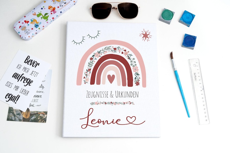 May include: A white rectangular folder featuring a printed design of a pink and red rainbow with floral accents, a heart, closed eyes, and a star. The text reads "ZEUGNISSE & URKUNDEN" and the name "Leonie" with a heart. A ruler is visible on the right.