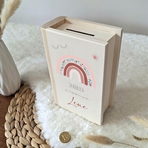 Op de afbeelding: Een houten spaarpot in de vorm van een boek, met een gleuf aan de bovenkant. De voorkant heeft een regenboogontwerp met de tekst "SPARBUCH" en de naam "Lene". De doos is licht van kleur met een natuurlijke houtnerf.
