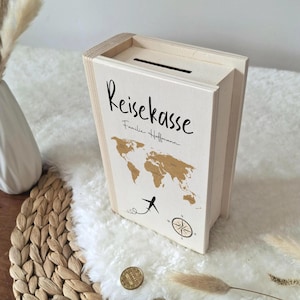 Può includere: Un salvadanaio da viaggio in legno chiaro con una fessura sulla parte superiore. La scatola presenta le parole "Reisekasse" e "Familie Hoffmann" in scrittura nera, insieme a una mappa del mondo dorata, una bussola e un aereo.