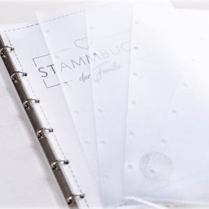 Könnte beinhalten: Ein weißer Ordner mit Metallringen, der halbtransparente Pergamentblätter enthält. Auf dem Deckblatt befinden sich ein Herzsymbol und der in Schwarz gedruckte Text STAMMBUCH der Familie. Die Seiten sind am linken Rand für die Ringmechanik gelocht.