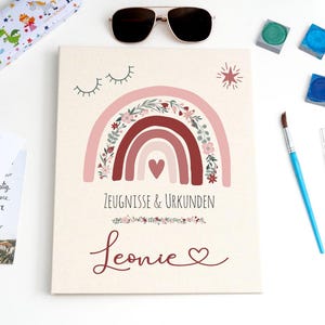 May include: A rectangular document folder with a cream background featuring a pink and red rainbow illustration with floral accents, a small heart, and closed eyes. The text reads Zeugnisse & Urkunden and the name Leonie in script font.