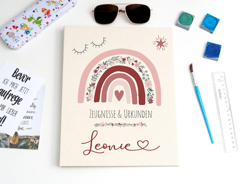 May include: A rectangular document folder with a cream background featuring a pink and red rainbow illustration with floral accents, a small heart, and closed eyes. The text reads Zeugnisse & Urkunden and the name Leonie in script font.