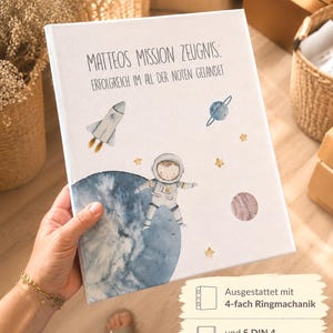 Peut inclure: Un classeur à couverture rigide blanche orné d'une illustration aquarelle d'un astronaute, d'une fusée, d'une planète bleue et d'étoiles jaunes. Le texte indique : MATTEOS MISSION ZEUGNIS: ERFOLGREICH IM ALL DER NOTEN GELANDET. Il possède 4 anneaux et 5 pochettes DIN A4.