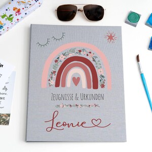 May include: A gray document folder featuring a central rainbow illustration with floral accents and a heart. The text reads Zeugnisse & Urkunden and the name Leonie in red script. Accessories include a pencil case, sunglasses, paint, and a ruler.
