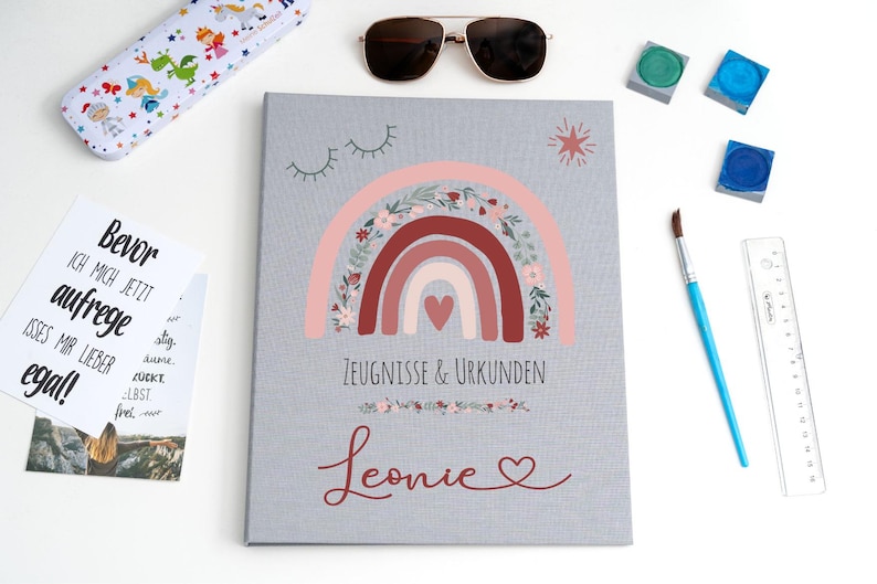 May include: A gray document folder featuring a central rainbow illustration with floral accents and a heart. The text reads Zeugnisse & Urkunden and the name Leonie in red script. Accessories include a pencil case, sunglasses, paint, and a ruler.