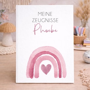 Op de afbeelding: Een wit notitieboek met de tekst "MEINE ZEUGNISSE Phoebe" in zwarte en roze letters. Een aquarel regenboogontwerp in roze tinten en een hartje onderaan. Het notitieboek ligt op een houten tafel.