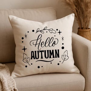 Può includere: Un cuscino quadrato color crema con le parole "Hello AUTUMN" in corsivo nero e lettere maiuscole. Il testo è circondato da un bordo decorativo di foglie, punti e stelle. Il cuscino è su un divano beige.