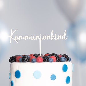 Könnte beinhalten: Weißer Kuchen, verziert mit blauen Punkten und mit frischen Beeren belegt. Ein weißer Kuchentopper lautet "Kommunionkind".