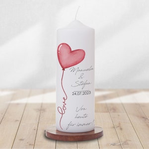 Könnte beinhalten: Weiße Kerze mit einem rosafarbenen herzförmigen Ballon und dem Text "Manuela & Stefan 24.07.2023 Von heute für immer!"  Love ist kursiv auf der Seite der Kerze geschrieben.